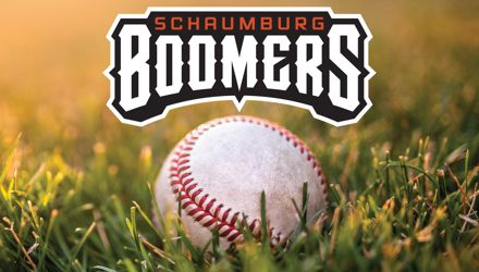 White baseball with red stitching lying in green grass. Golden sunlight filtering from above. Schaumburg Boomers logo in the top, center of the image.