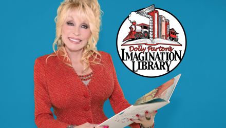 Dolly Parton, smiling, dressed in a red blazer, holding an open children's book in front of a blue background. The Imagination Library logo in the upper central portion of the image.